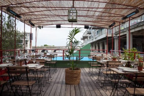 Le patio extérieur de LE RÊVE FLOTTANT est composé de tables et de chaises en bois, d'une grande plante en pot au centre, d'un toit en bambou, de guirlandes lumineuses et d'une végétation luxuriante, le tout créant un cadre confortable en plein air avec vue sur le paysage urbain.