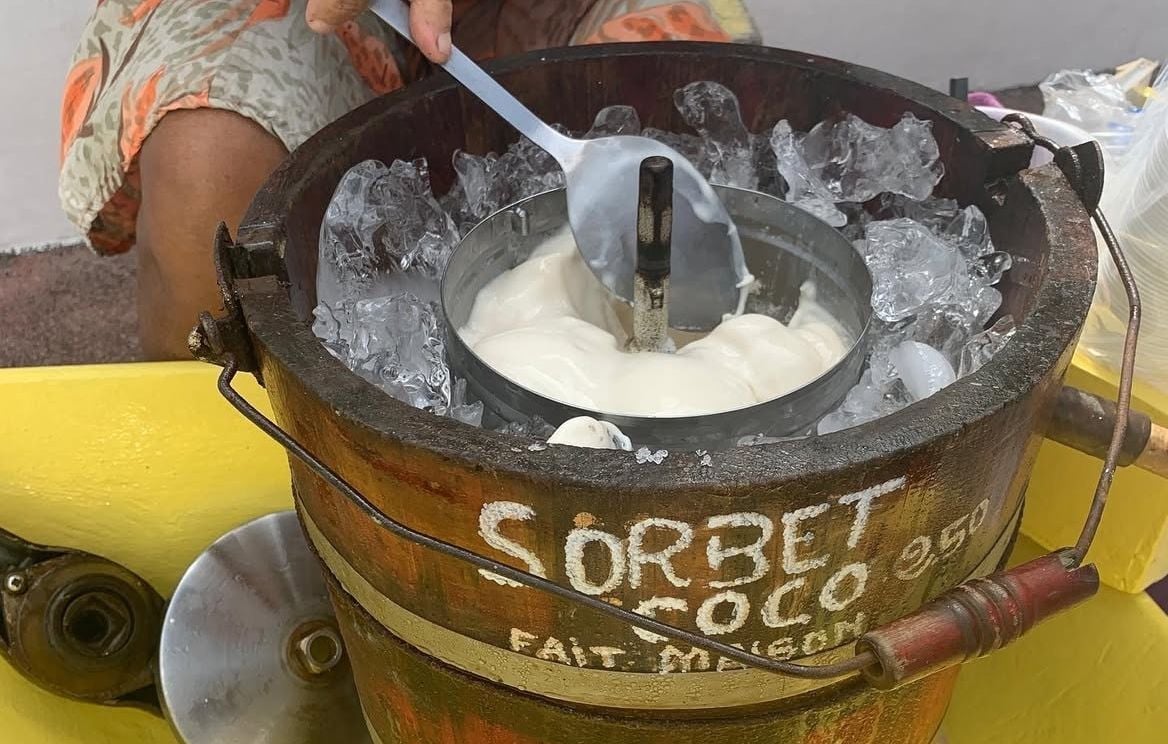 Une personne remue un sorbet crémeux à la noix de coco dans un récipient métallique entouré de glace à l'intérieur d'un seau en bois étiqueté SORBET COCO 950 FAIT MAISON.