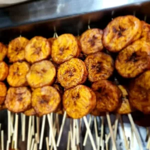BROCHETTES DE BANANES PLANTAIN : Tranches de plantain fraîchement cuites et dorées sur des brochettes, disposées en rangées. Commande minimum : 10 brochettes.