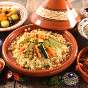 Profitez de l'ANIMATION COUSCOUS PARTY : un couscous marocain traditionnel avec des pois chiches, des carottes et des courgettes, servi dans un tajine en terre cuite avec un assortiment de légumes et de sauces sur une table en bois rustique.