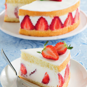 Une tranche de FRAISIER (32 parts) avec des couches de génoise, des fraises fraîches et de la crème est posée sur une assiette à côté du gâteau entier garni de fraises et de menthe. Une fourchette est posée à côté de la tranche.