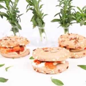 Un plateau de MINI BAGEL LEGUMES DU SOLEIL (Plateau de 20 pièces), garni de fromage et de tomates, est présenté sur une surface blanche avec des feuilles vertes, tandis que des bocaux en verre à l'arrière-plan contiennent des brins d'herbes fraîches.