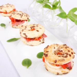 Les MINI BAGELS PASTRAMI (Plateau de 20 pièces) sont disposés en rang sur une assiette blanche, garnis de pastrami, de fromage et de poivrons rouges, décorés de feuilles vertes et de petits vases avec des tiges vertes en arrière-plan.