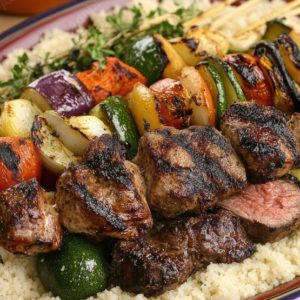 GRILLADES DE COUSCOUS : Brochettes de bœuf grillé avec tomates, oignons, poivrons et courgettes servies sur un couscous moelleux, agrémenté d'herbes fraîches.