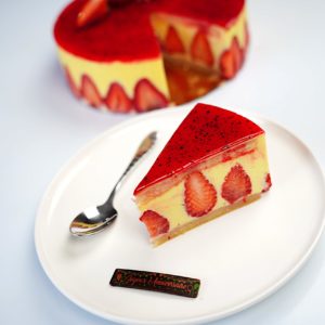 Une tranche de FRAISIER (24 parts) avec des morceaux de fraises visibles est posée sur une assiette blanche à côté d'une cuillère en argent, tandis que le gâteau entier avec un dessus glacé rouge et des couches de mousse jaune apparaît à l'arrière-plan.