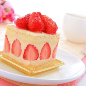 Une tranche de FRAISIER (12 parts) garnie de fraises fraîches repose sur une assiette blanche, accompagnée d'une tasse et de fleurs roses en arrière-plan.