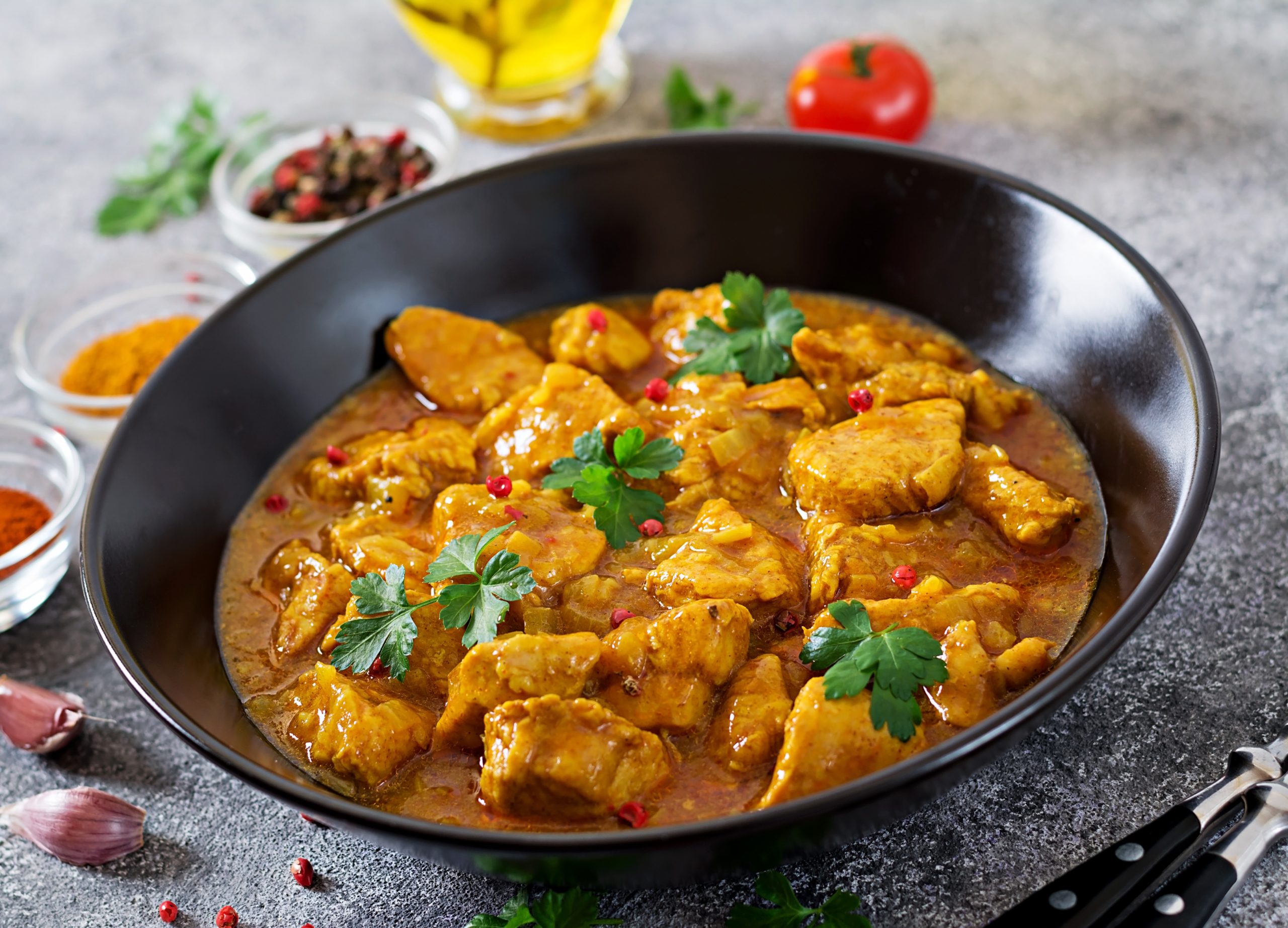 Un bol noir de COLOMBO DE POULET (300g la part) contenant des morceaux de poulet au curry dans une riche sauce jaune, agrémentée de coriandre fraîche et de poivre rouge. En arrière-plan flou, une tomate, des épices et un récipient à huile en verre.