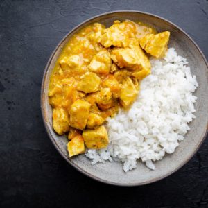 Une portion de COLOMBO DE POULET (300g la part) avec du riz blanc d'un côté et du curry de poulet jaune avec des morceaux de poulet et de la sauce de l'autre, présentée sur une surface foncée.