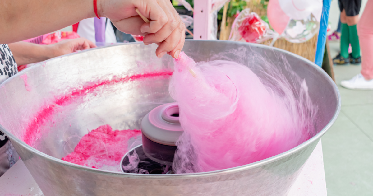 Une personne fait tourner une barbe à papa rose autour d'un bâton à l'intérieur d'une grande machine métallique, formant ainsi une friandise duveteuse lors d'un événement en plein air.