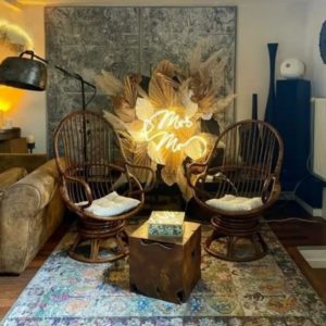 Un salon confortable avec LE LOFT ART FEELING (93170) BAGNOLET, avec deux chaises rondes en osier, des coussins, une table rustique en bois et une enseigne au néon Mrs & Mrs au milieu de feuilles séchées et d'herbe de la pampa pour une ambiance chaleureuse et invitante.