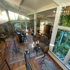 Chambre spacieuse et lumineuse à LE LOFT ART FEELING (93170) BAGNOLET avec parquet, grandes fenêtres, tapis, plantes et pupitres. La lumière naturelle remplit l'espace confortable avec de la verdure visible à l'extérieur. Le logo de Magnolia For Event est affiché dans le coin.