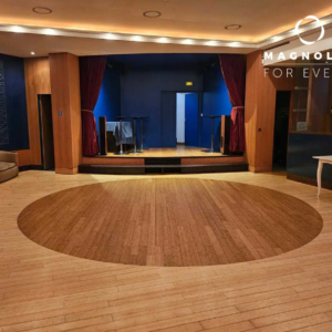 Une salle d'événement spacieuse avec un sol en bois clair, un centre circulaire plus sombre, une petite scène avec des rideaux rouges, des murs bleus, un mobilier minimal. L'inscription "L'AUDIENCE (92130) ISSY LES MOULINEAUX" figure dans le coin supérieur droit.