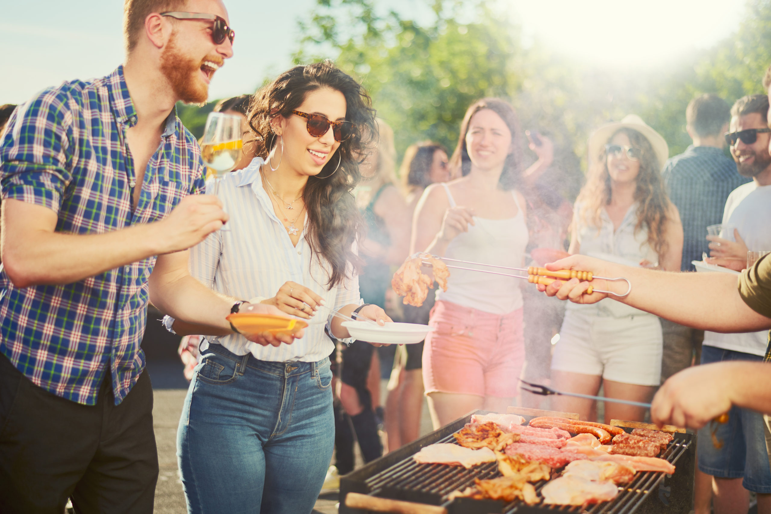 Un groupe de personnes souriant et socialisant lors d'un barbecue en plein air, avec de la nourriture grillée et servie. Le soleil brille et tout le monde semble profiter de cette rencontre décontractée.