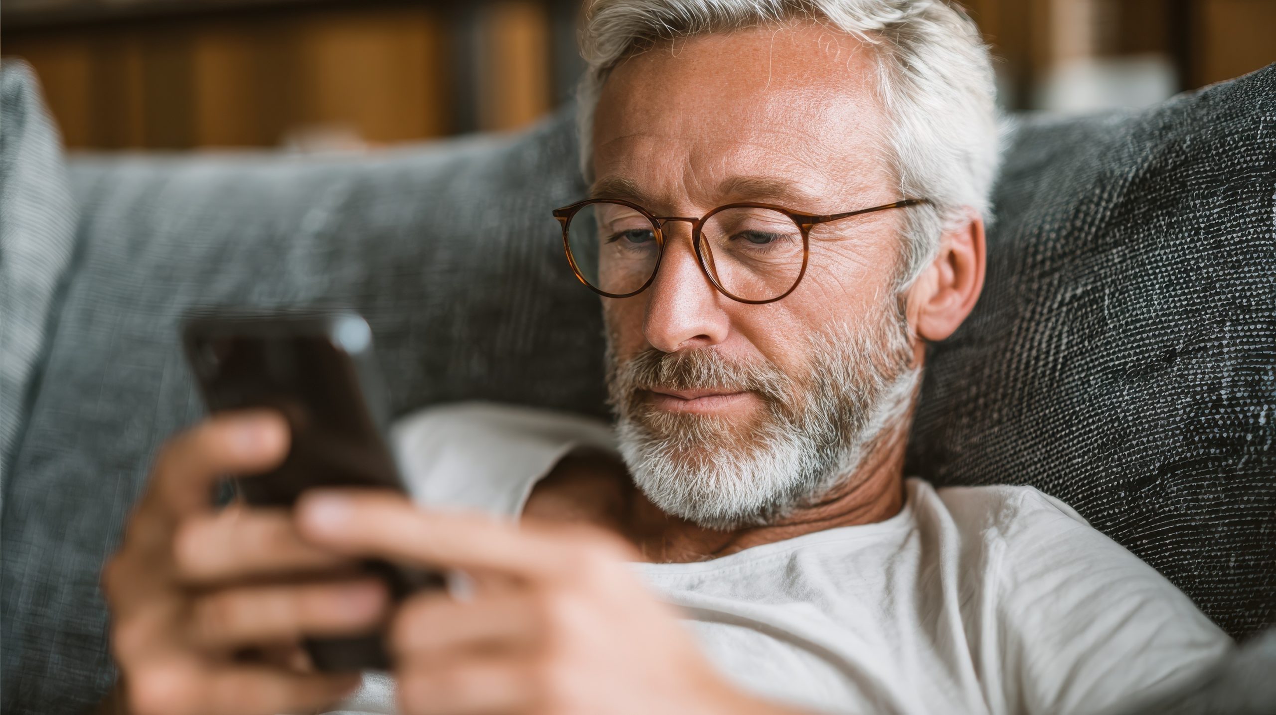 Un homme d'âge moyen avec des cheveux gris, une barbe et des lunettes est allongé sur un canapé, vêtu d'une chemise blanche, et regarde un smartphone qu'il tient à deux mains.