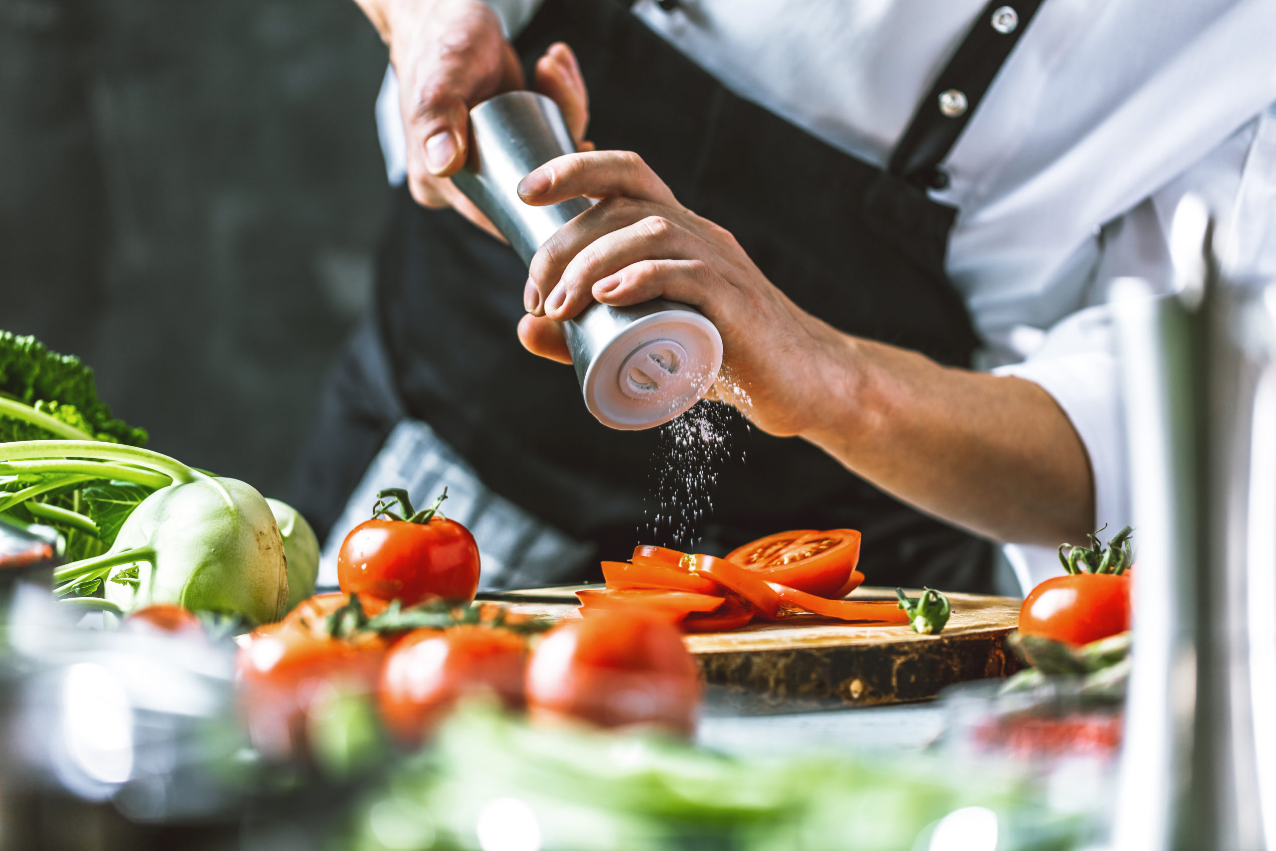 Une personne vêtue d'un tablier noir saupoudre du sel provenant d'un broyeur métallique sur des tranches de tomates posées sur une planche à découper en bois, entourée de légumes frais dans une cuisine.