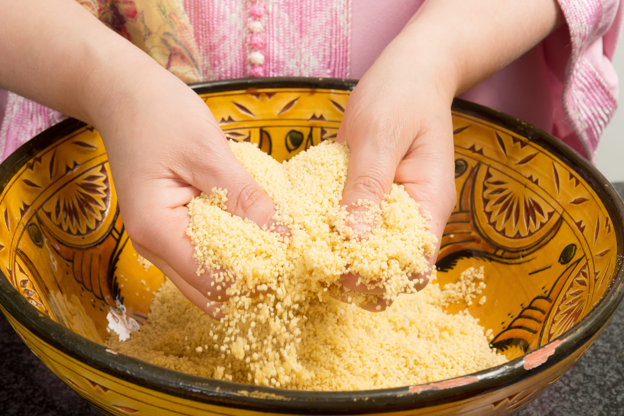 Mains mélangeant et gonflant du couscous jaune dans un bol en céramique orné de motifs, quelques grains de couscous tombant entre les doigts. La personne porte un vêtement rose.