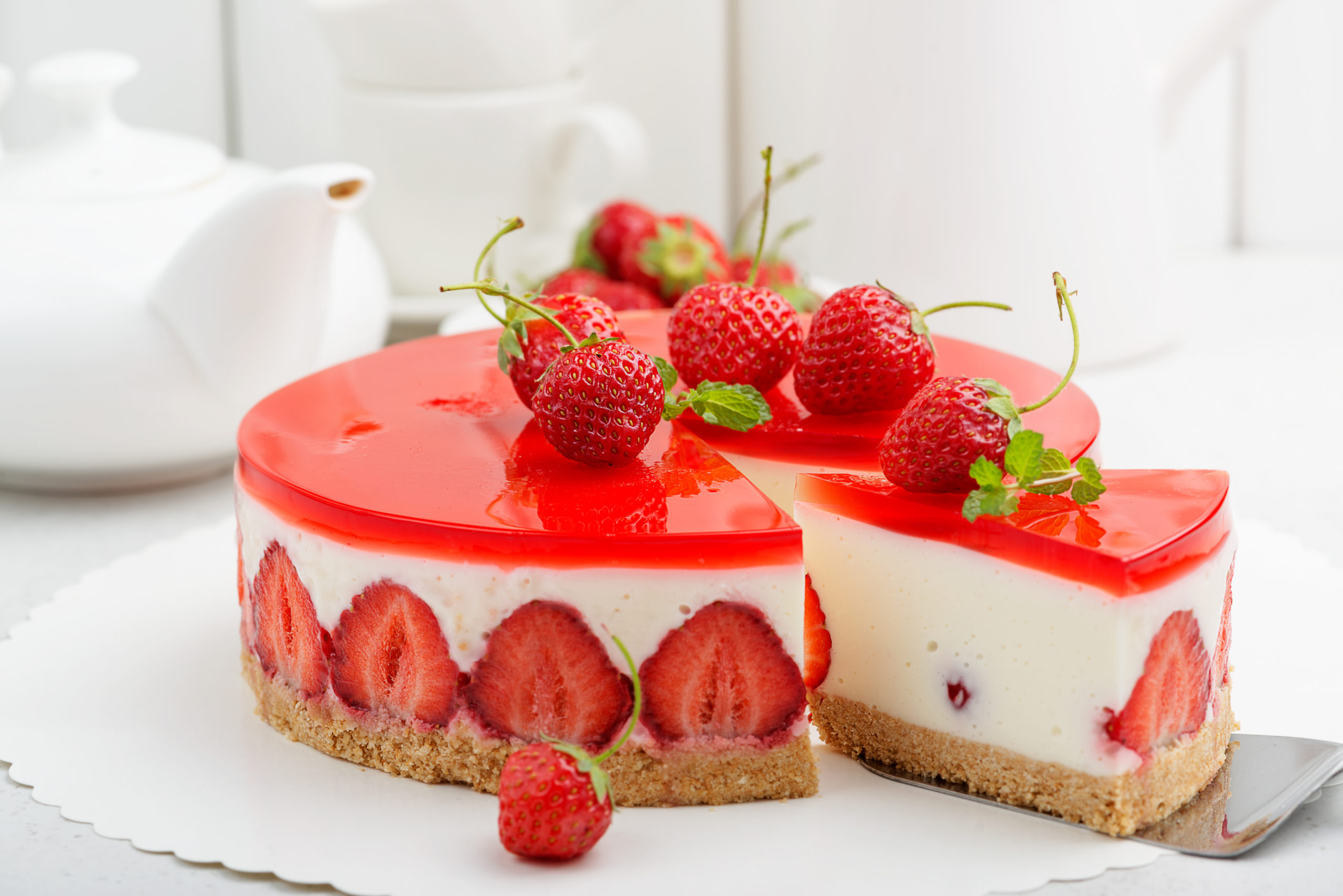 Gâteau au fromage à la fraise, nappé de gelée rouge transparente et de fraises fraîches, avec une base biscuitée et des fraises entières à l'intérieur. Une tranche est soulevée et une théière est visible à l'arrière-plan.