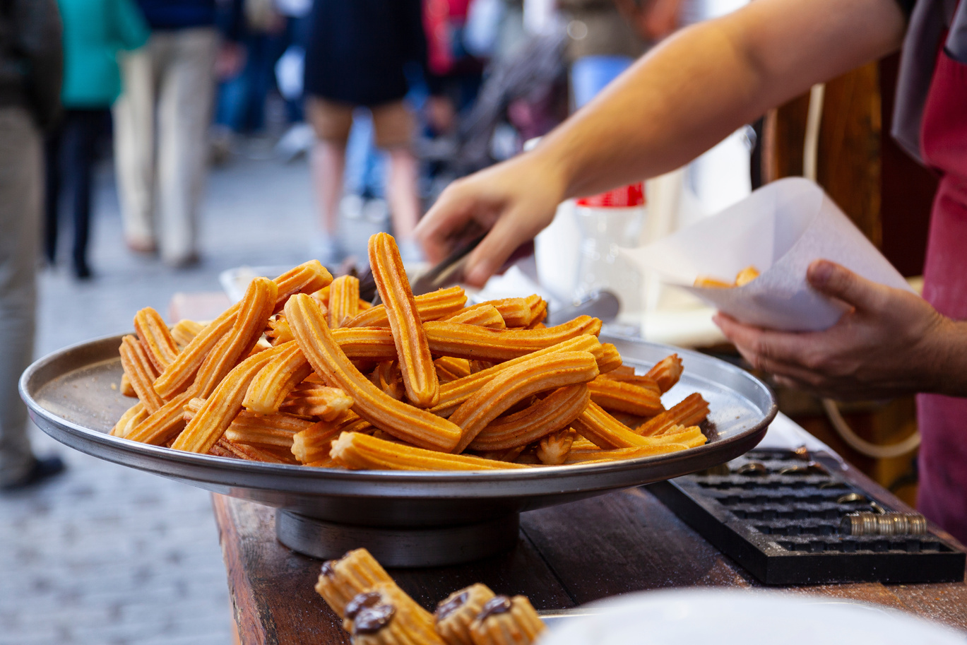 Un grand plateau rempli de churros est posé sur une table en bois sur un marché en plein air. Une personne utilise des pinces pour prendre des churros et les placer dans un porte-papier, avec des personnes floues à l'arrière-plan.