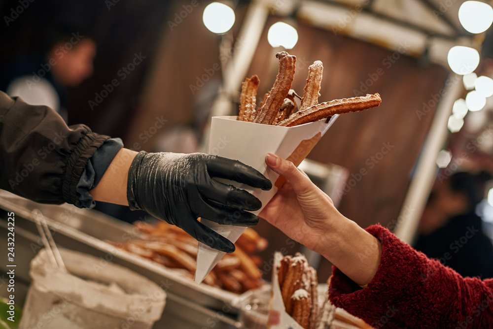 Un vendeur portant un gant noir tend un cornet en papier rempli de churros à un client vêtu d'un pull-over rouge sur un étal de marché, avec des lumières vives floues en arrière-plan.