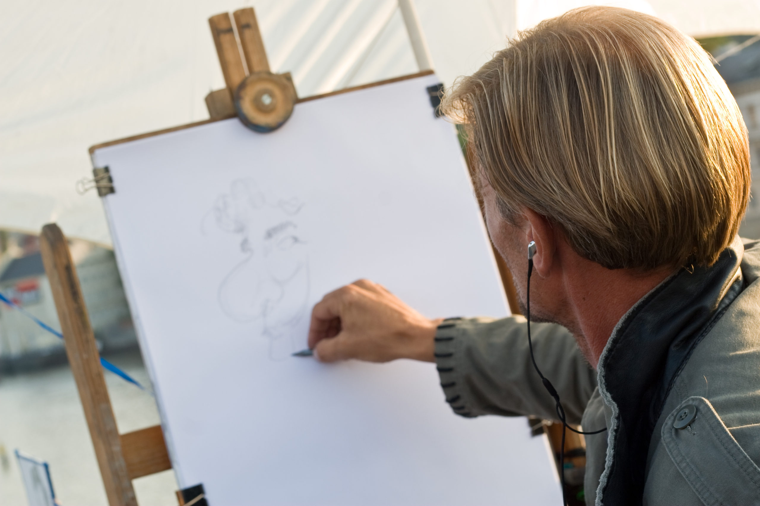 Une personne aux cheveux blonds esquisse une caricature sur une grande feuille blanche fixée à un chevalet en plein air. L'artiste semble concentré, il porte une veste légère et écoute des écouteurs.