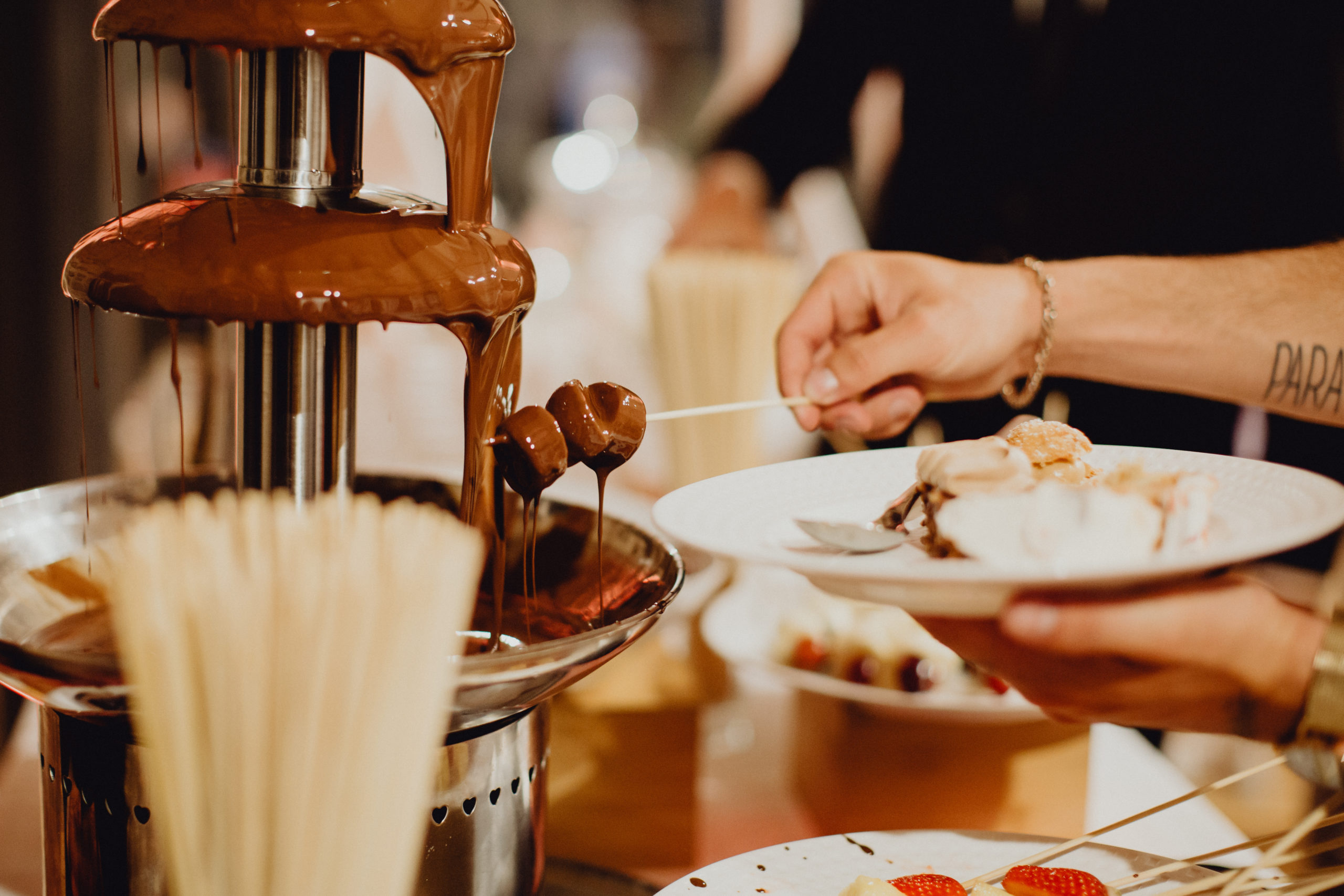 Une personne trempe des brochettes dans une fontaine de chocolat qui coule sur une table de desserts, en tenant une assiette contenant diverses friandises. Des bâtonnets et des bols contenant d'autres friandises sont visibles à l'arrière-plan.