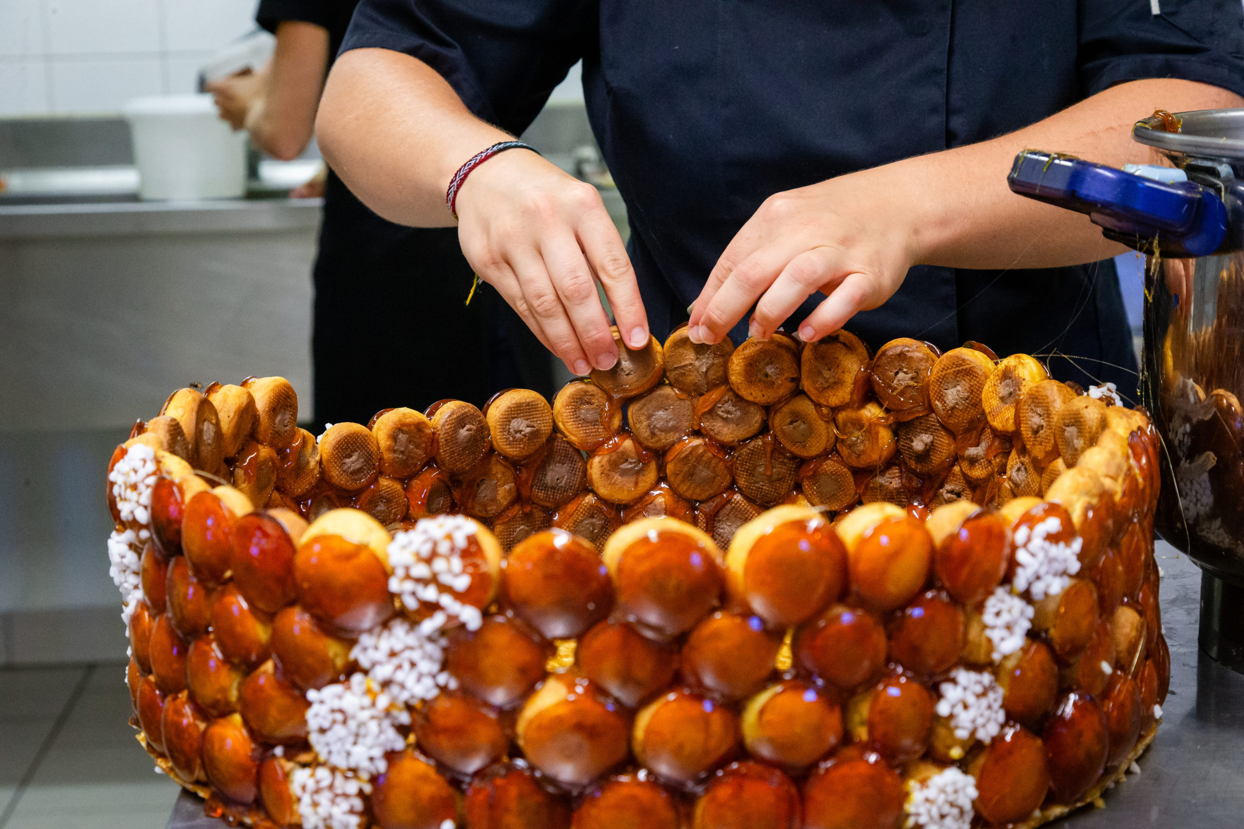 Dans une cuisine professionnelle, une personne vêtue d'une veste de chef sombre assemble avec soin un grand croquembouche rond composé de choux à la crème enrobés de caramel.