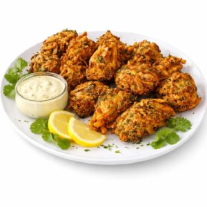 BEIGNETS PAKORA (Minimum de commande 10 pièces) : des beignets dorés servis sur une assiette blanche, accompagnés d'herbes fraîches, de tranches de citron, de coriandre et d'une sauce crémeuse en accompagnement.