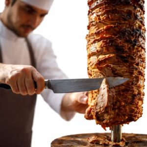 Un chef en uniforme blanc découpe de la viande cuite dans une rôtissoire verticale pour préparer le POULET CHAWARMA (Plat - Min de commande 5 plats de 300g/Pers) sur un fond blanc.