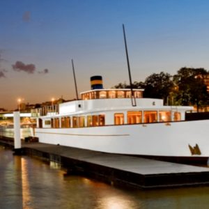 LE YACHT CLIPPER (75015), un grand yacht blanc avec un entonnoir bleu et jaune, est à quai au coucher du soleil, illuminé sur une eau calme qui reflète les lumières de la ville, avec des arbres et des bâtiments en arrière-plan.