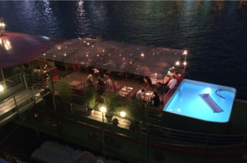 La nuit, LE RÊVE FLOTTANT est ancré sur la rivière avec une salle à manger extérieure, des guirlandes lumineuses et un bassin lumineux avec une chaise longue flottante où les clients profitent du dîner et de l'atmosphère tranquille.