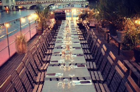Le LE RÊVE FLOTTANT propose un dîner du soir au bord de l'eau sur un pont, avec une longue table garnie de verres et de chaises vides, entourée de guirlandes lumineuses, de plantes et de vues sur le pont et l'eau.