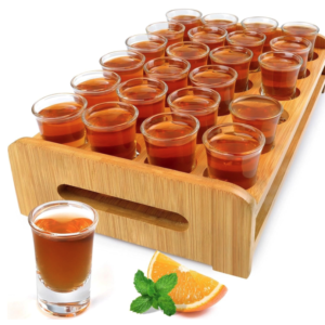 Le PLATEAU DE VERRE À TI PUNCH présente 24 verres à shot remplis sur un plateau en bois, avec un seul verre, une tranche d'orange et un brin de menthe disposés devant sur un fond blanc.