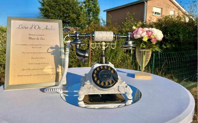 Un téléphone rotatif vintage, un panneau encadré et un vase de fleurs roses et blanches sont disposés sur une table blanche à l'extérieur, avec de la verdure et une maison en arrière-plan.