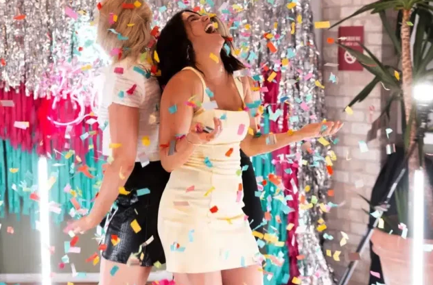 Deux femmes se tiennent dos à dos, riant joyeusement tandis que des confettis colorés tombent autour d'elles. Elles sont à l'intérieur, avec des décorations festives et des rideaux de guirlandes chatoyants en arrière-plan.