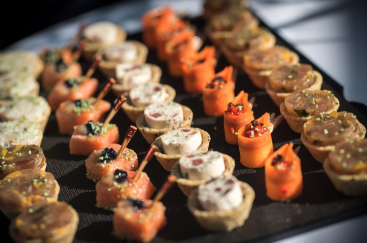 Gros plan sur un assortiment d'amuse-gueules gastronomiques, dont des tartelettes, des bouchées au saumon fumé et des rouleaux de carottes, disposés en rangées sur un plateau noir.