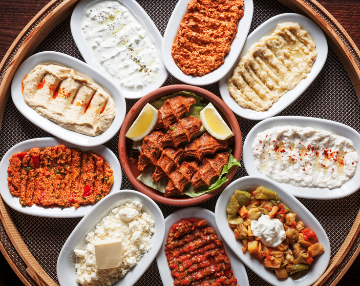 Un plateau rond contient une variété de plats meze turcs dans de petites assiettes ovales, notamment du houmous, des dips au yaourt, des salades de légumes et une assiette centrale de tranches de viande épicées garnies de laitue et de quartiers de citron.