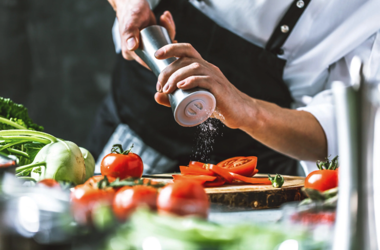 Une personne en uniforme de chef broie du sel sur des tranches de tomates sur une planche à découper, entourée de légumes frais tels que des tomates et des légumes verts dans une cuisine.