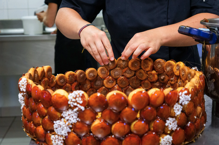 Une personne en uniforme sombre de chef cuisinier dispose des choux à la crème glacés au caramel en une grande structure circulaire, peut-être un croquembouche, sur un comptoir de cuisine avec des carreaux et des ustensiles blancs à l'arrière-plan.