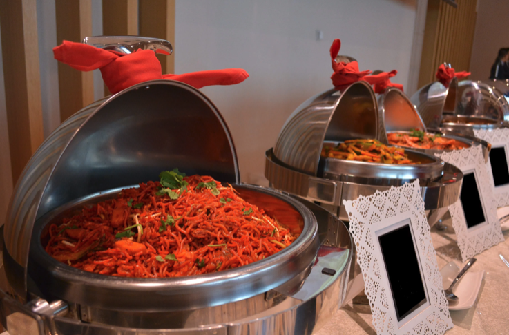 Trois chafing dishes remplis d'aliments variés, dont un plat de nouilles garni de coriandre, sont disposés sur une table de buffet. Chaque plat est recouvert d'un couvercle et des cartes de menu vierges sont placées devant.