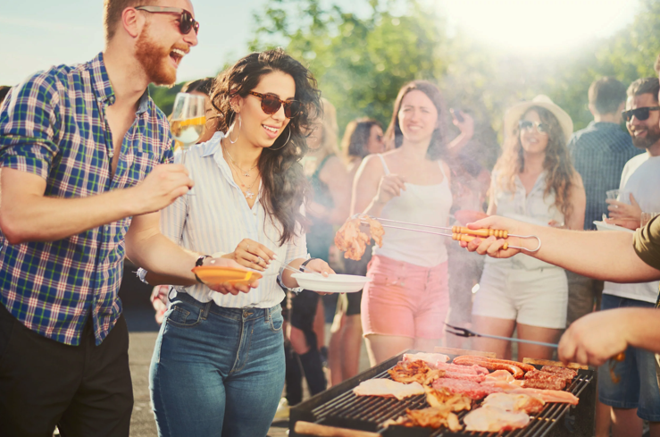 Un groupe de personnes participant à une fête barbecue en plein air, souriant et riant. Deux personnes se tiennent près d'un gril avec de la nourriture, l'une tenant des pinces et faisant griller de la viande tandis que les autres tiennent des assiettes et des boissons. Le soleil brille de mille feux.