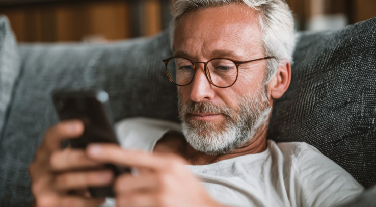 Un homme d'âge moyen, aux cheveux gris, à la barbe et aux lunettes, est allongé sur un canapé. Il porte une chemise blanche et regarde son smartphone, semblant détendu et concentré.