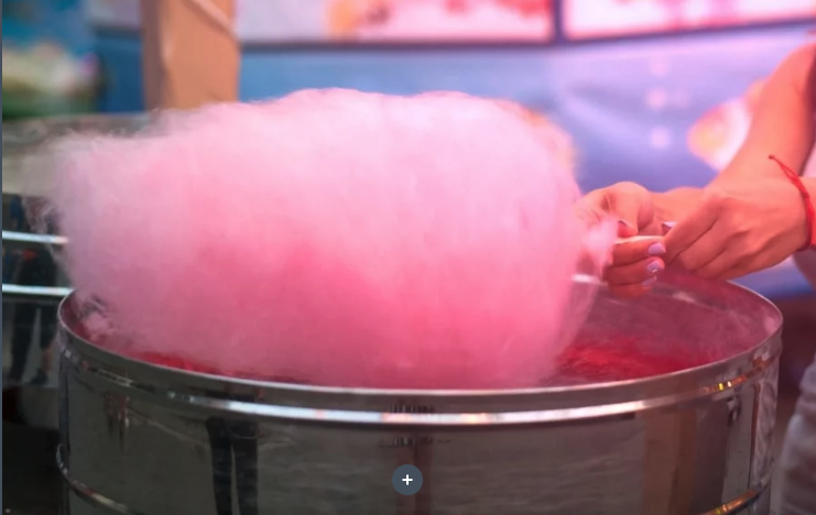 Une personne fait tourner une barbe à papa rose sur un bâton au-dessus d'une grande machine à barbe à papa en métal lors d'une foire ou d'un événement.