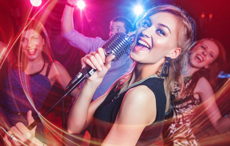 Une jeune femme chante dans un microphone vintage lors d'une fête animée, le sourire aux lèvres. Des lumières colorées et des personnes floues dansant en arrière-plan créent une atmosphère énergique et festive.