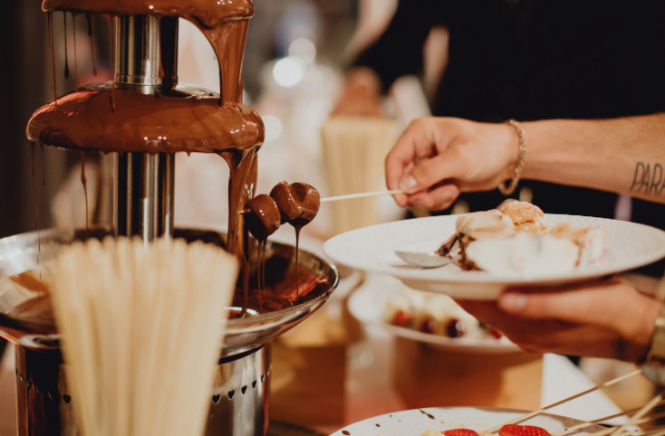 Une personne trempe des brochettes dans une fontaine de chocolat qui coule, en tenant une assiette blanche avec des desserts assortis. D'autres brochettes et assiettes de nourriture sont visibles sur la table à proximité.