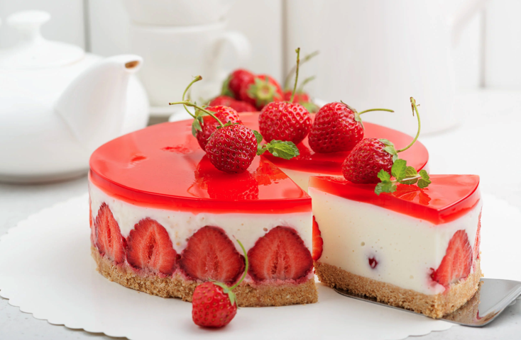 Un cheesecake rond aux fraises avec une couche de gelée rouge brillante, des fraises entières à l'intérieur et sur le dessus, et une base en biscuit. Une tranche est servie, montrant la garniture crémeuse et les fraises fraîches.