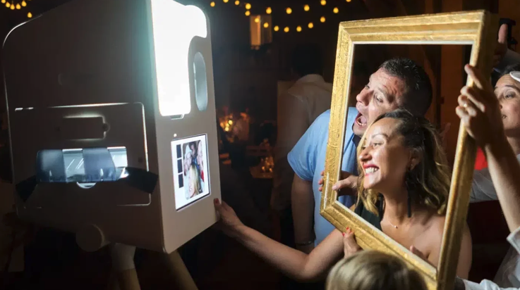 Un couple pose et sourit devant la caméra d'un photomaton tout en tenant un cadre photo vide autour de leur visage lors d'un événement festif avec des guirlandes lumineuses en arrière-plan.