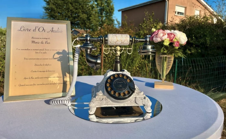 Un téléphone à cadran vintage est posé sur une table ronde avec une enseigne à cadre doré et un bouquet de fleurs roses et blanches dans un vase. La table se trouve à l'extérieur, près d'une maison et d'arbres, sous un ciel bleu clair.
