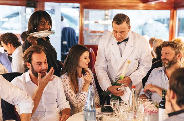 Un serveur en veste blanche sert un groupe de quatre personnes assises à une table ronde de restaurant, tandis qu'un autre serveur apporte des assiettes. Le groupe est engagé et souriant, entouré de verres et de bouteilles.