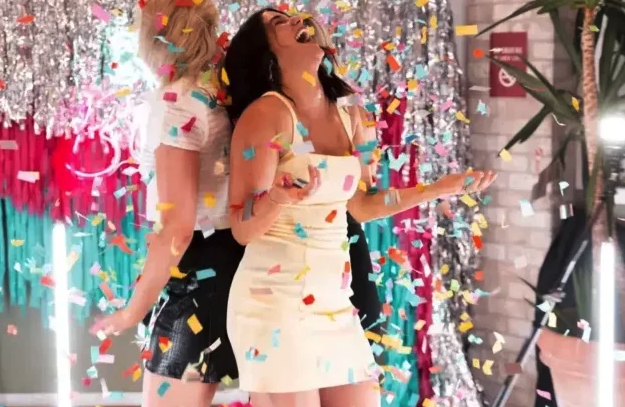 Deux femmes se tiennent dos à dos et rient joyeusement tandis que des confettis colorés tombent autour d'elles. Elles sont entourées de décorations festives, dont des serpentins argentés et roses brillants.
