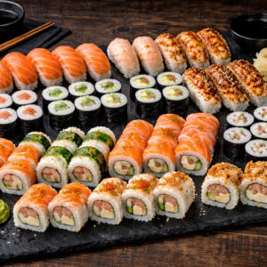 Le PLATEAU DE SUSHIS 75 pièces présente un assortiment de rouleaux de sushi, de nigiri et de maki disposés avec art sur un grand plateau d'ardoise avec des bols de wasabi, de gingembre mariné et de sauce soja posés sur une table en bois sombre.